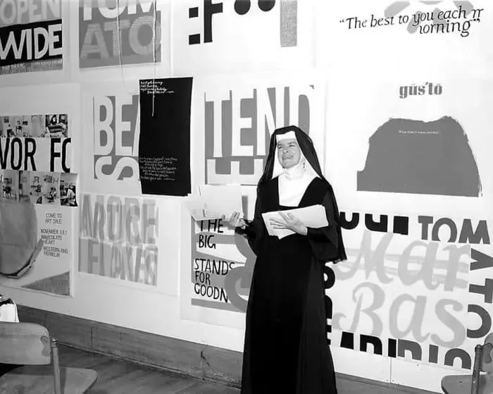 Fotografia en blanc i negre: Corita, amb hàbit, dreta i somrient davant d'una paret coberta de serigrafies amb tipografia gran, amb paraules com OPEN WIDE, TOMATO o THE BIG STANDS FOR GOODNESS.