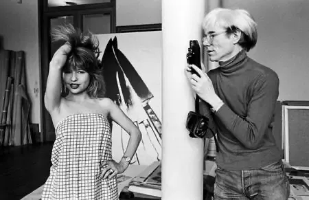 Fotografia en blanc i negre: Andy Warhol, amb cabell clar i coll alt fosc, apunta una càmera cap a una dona que posa dreta davant seu, en un estudi amb quadres recolzats a la paret.